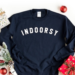 Puede incluir: Sudadera azul marino con el texto "INDOORSY" en letras blancas. La sudadera est&aacute; rodeada de decoraciones navide&ntilde;as, incluyendo un regalo envuelto, ramas de pino y adornos rojos.