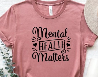 May include: A dusty rose t-shirt with black text that reads "Mental Health Matters" with a heart symbol on either side of the word "Health".