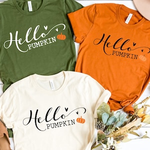 May include: Three t-shirts in olive green, orange, and cream colors, each with the text "Hello Pumpkin" in a script font, a heart, and a small pumpkin graphic. The shirts are displayed with fall-themed decorations.