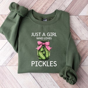 Puede incluir: Una sudadera verde con el texto "Just a girl who loves pickles" y una ilustraci&oacute;n de dibujos animados de un frasco de pepinillos con un lazo rosa.