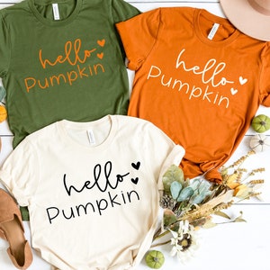 May include: Three t-shirts with the text "hello pumpkin" in different colors: olive green, orange, and cream. The shirts are laid out on a white wooden surface with fall decor.