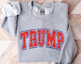 May include: Gray sweatshirt with a red and blue "TRUMP" graphic in a collegiate font. The letters are outlined in blue and filled with red glitter.