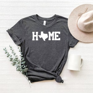 May include: A dark gray t-shirt with a white outline of the state of Texas and the word "HOME" printed on the front. The shirt is laid out on a white textured surface with a white mug and a beige hat.