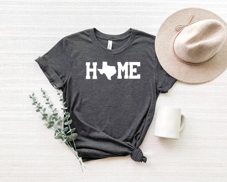 May include: A dark gray t-shirt with a white outline of the state of Texas and the word "HOME" printed on the front. The shirt is laid out on a white textured surface with a white mug and a beige hat.