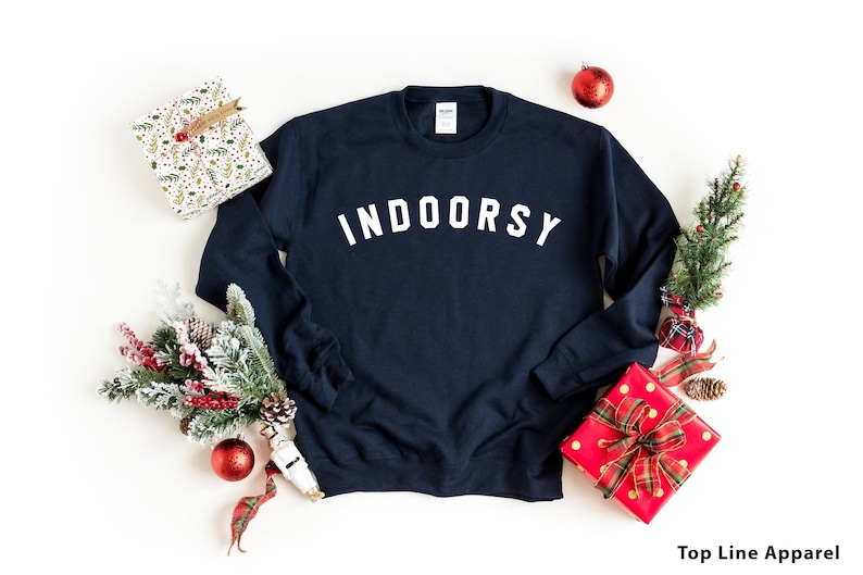 Puede incluir: Sudadera azul marino con el texto "INDOORSY" en letras blancas. La sudadera est&aacute; rodeada de decoraciones navide&ntilde;as, incluyendo un regalo envuelto, ramas de pino y adornos rojos.