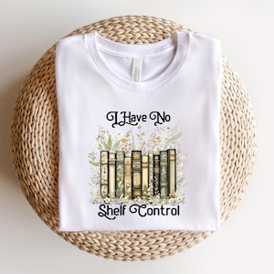 May include: White t-shirt featuring the text "I Have No Shelf Control" in black script. The design includes a watercolor illustration of a stack of books surrounded by floral elements. The shirt is displayed on a woven, circular surface.
