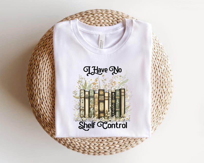 May include: White t-shirt featuring the text "I Have No Shelf Control" in black script. The design includes a watercolor illustration of a stack of books surrounded by floral elements. The shirt is displayed on a woven, circular surface.