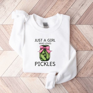 Puede incluir: Sudadera blanca con el texto "Just a girl who loves pickles" y un dibujo de un tarro de pepinillos con un lazo rosa.