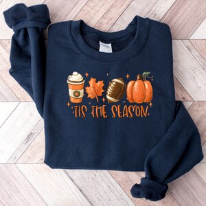K&ouml;nnte beinhalten: Navyblauer Sweatshirt mit einem grafischen Design aus einer Kaffeetasse, einem Blatt, einem Football und einem K&uuml;rbis. Der Text "Tis the Season" ist unter dem Grafikdruck.