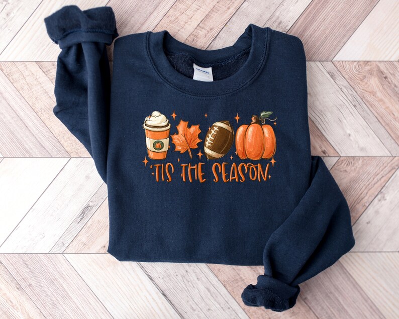 K&ouml;nnte beinhalten: Navyblauer Sweatshirt mit einem grafischen Design aus einer Kaffeetasse, einem Blatt, einem Football und einem K&uuml;rbis. Der Text "Tis the Season" ist unter dem Grafikdruck.