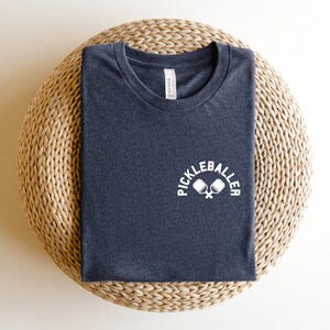May include: A navy blue t-shirt featuring the white text "PICKLEBALLER" above a graphic of crossed pickleball paddles. The shirt is folded on a circular woven mat. The t-shirt is a dark blue colour.