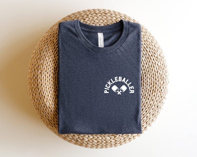 May include: A navy blue t-shirt featuring the white text "PICKLEBALLER" above a graphic of crossed pickleball paddles. The shirt is folded on a circular woven mat. The t-shirt is a dark blue colour.