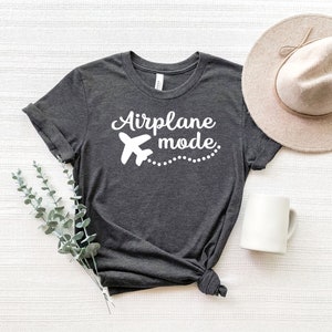 May include: A dark grey t-shirt with the text "Airplane mode" in white lettering. A white airplane icon is next to the text. The shirt is laid flat on a white surface with a brown hat and a white mug.