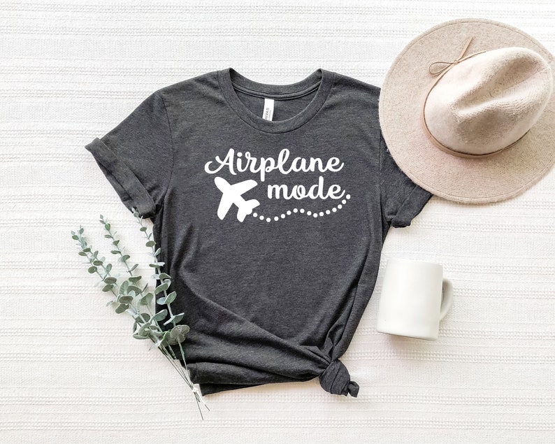 May include: A dark grey t-shirt with the text "Airplane mode" in white lettering. A white airplane icon is next to the text. The shirt is laid flat on a white surface with a brown hat and a white mug.
