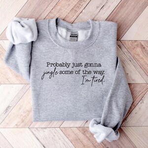 Puede incluir: Una sudadera gris jaspeada con el texto "Probably just gonna jingle some of the way. I'm tired." en escritura negra. La sudadera est&aacute; doblada, con las mangas visibles.