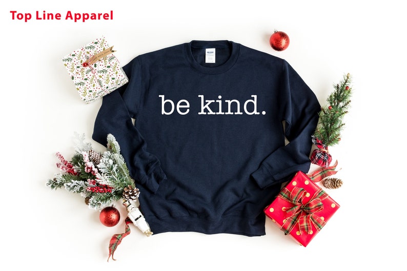May include: A navy blue sweatshirt with the text "be kind." printed in white on the front. The sweatshirt is surrounded by Christmas decorations, including a small Christmas tree, red ornaments, and wrapped gifts.