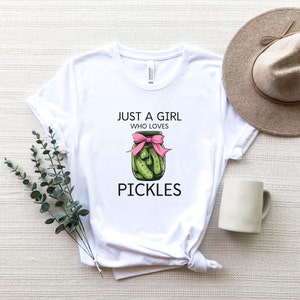 Puede incluir: Camiseta blanca con el texto "Just a girl who loves pickles" y un dibujo de un frasco de pepinillos con una cinta rosa.