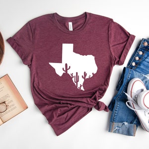 May include: A maroon t-shirt with a white outline of the state of Texas with cactus silhouettes inside. The t-shirt is laid flat on a white surface with a pair of blue jeans and white sneakers.