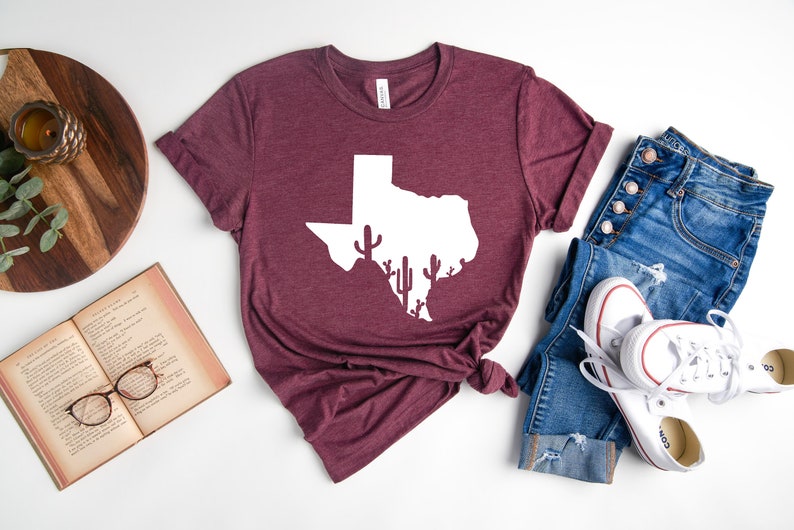 May include: A maroon t-shirt with a white outline of the state of Texas with cactus silhouettes inside. The t-shirt is laid flat on a white surface with a pair of blue jeans and white sneakers.
