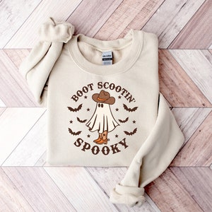 May include: A beige sweatshirt with a graphic print of a ghost wearing a cowboy hat and boots. The text "Boot Scootin' Spooky" is printed around the ghost.