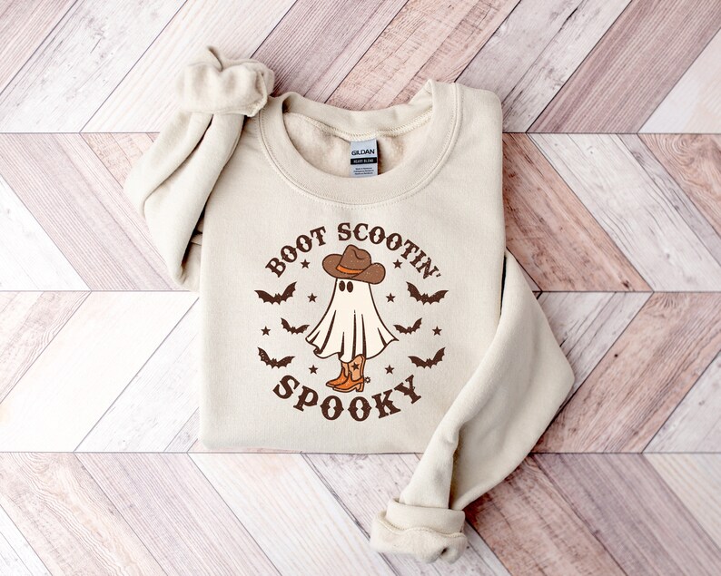 May include: A beige sweatshirt with a graphic print of a ghost wearing a cowboy hat and boots. The text "Boot Scootin' Spooky" is printed around the ghost.