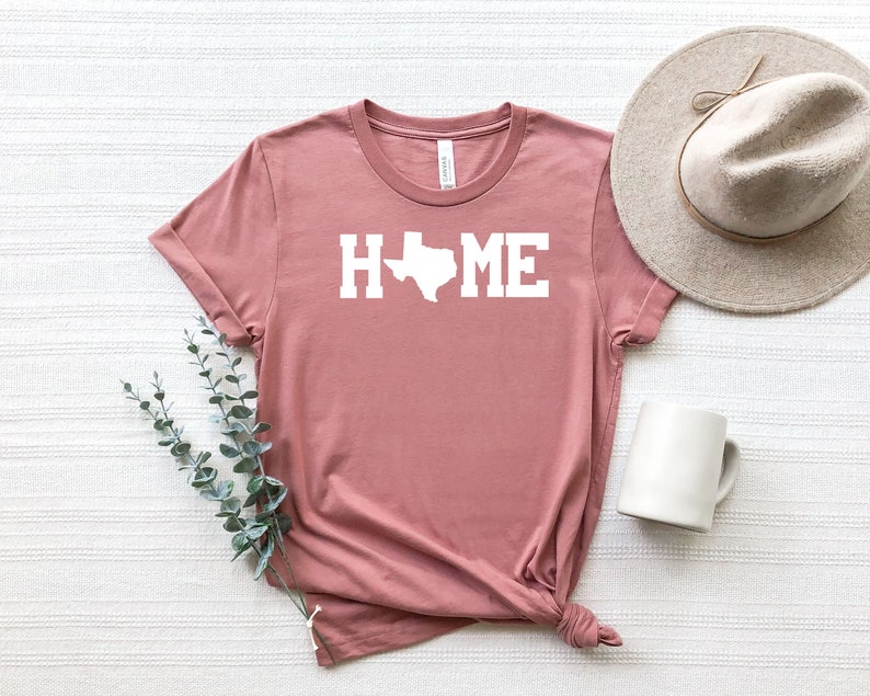 May include: A dusty rose t-shirt with the word "HOME" in white text with the outline of the state of Texas replacing the "O".