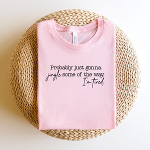 Puede incluir: Una camiseta rosa claro con el texto negro "Probably just gonna jingle some of the way. I'm tired." La camiseta est&aacute; doblada y colocada sobre una superficie circular tejida.