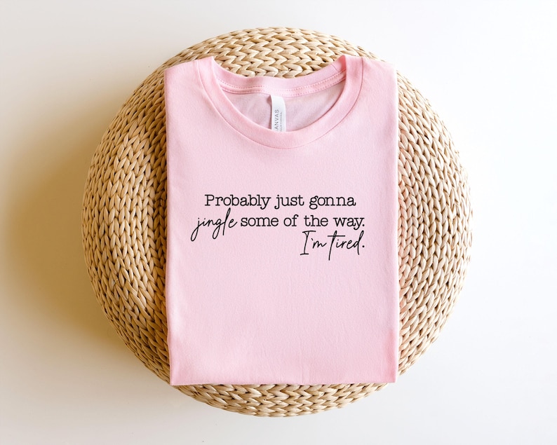 Puede incluir: Una camiseta rosa claro con el texto negro "Probably just gonna jingle some of the way. I'm tired." La camiseta est&aacute; doblada y colocada sobre una superficie circular tejida.