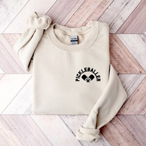 May include: A cream-coloured sweatshirt featuring the text "PICKLEBALLER" above a graphic of two pickleball paddles. The sweatshirt has a crew neck and long sleeves with ribbed cuffs.