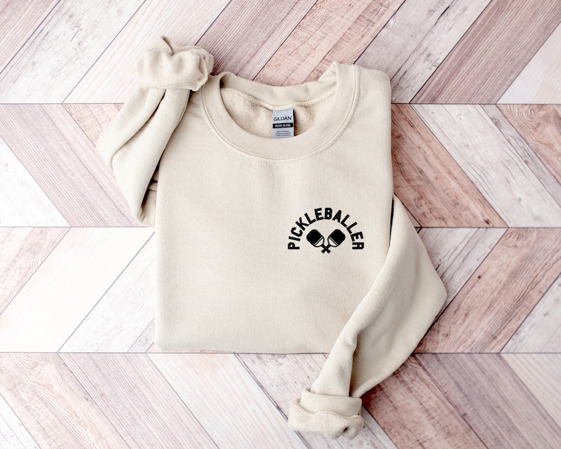 May include: A cream-coloured sweatshirt featuring the text "PICKLEBALLER" above a graphic of two pickleball paddles. The sweatshirt has a crew neck and long sleeves with ribbed cuffs.