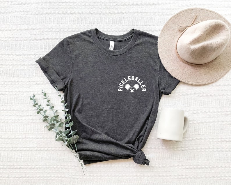 May include: Dark grey t-shirt with the text "PICKLEBALLER" in white above crossed pickleball paddles. A beige hat and white mug are next to the shirt. A sprig of green leaves is on the left.