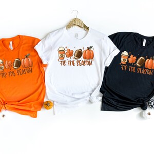 K&ouml;nnte beinhalten: Drei T-Shirts mit einem Herbst-Motiv, das eine Kaffeetasse, Bl&auml;tter, einen Football und einen K&uuml;rbis zeigt. Der Text auf den T-Shirts lautet "'Tis the season".