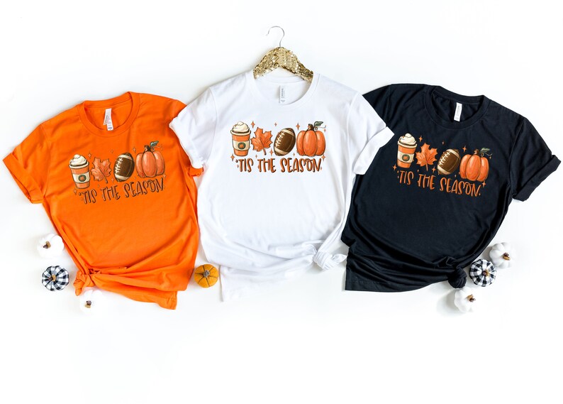 K&ouml;nnte beinhalten: Drei T-Shirts mit einem Herbst-Motiv, das eine Kaffeetasse, Bl&auml;tter, einen Football und einen K&uuml;rbis zeigt. Der Text auf den T-Shirts lautet "'Tis the season".