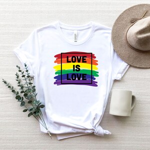 May include: White t-shirt with the text "LOVE IS LOVE" in black, set against a rainbow-colored brushstroke design. The shirt is styled with a neutral-toned hat and a white mug. A sprig of greenery is placed to the left of the shirt.