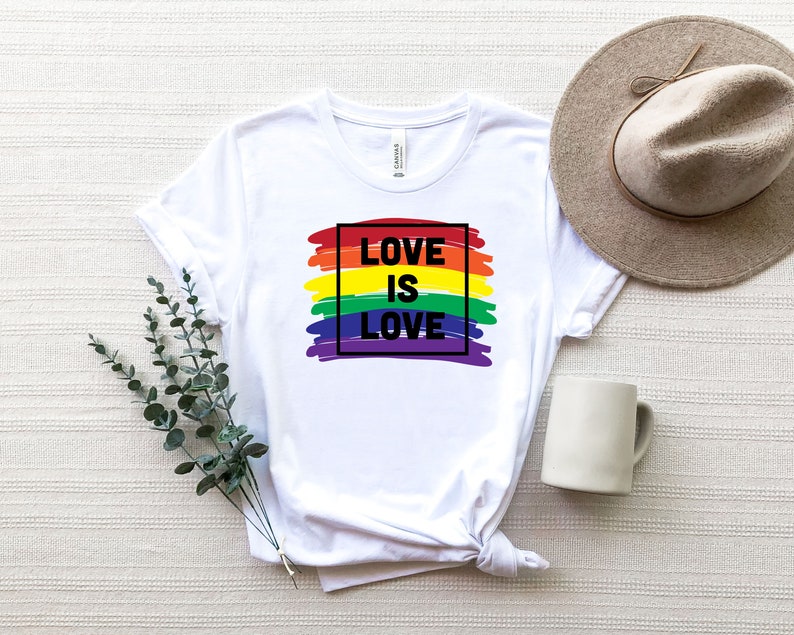 May include: White t-shirt with the text "LOVE IS LOVE" in black, set against a rainbow-colored brushstroke design. The shirt is styled with a neutral-toned hat and a white mug. A sprig of greenery is placed to the left of the shirt.