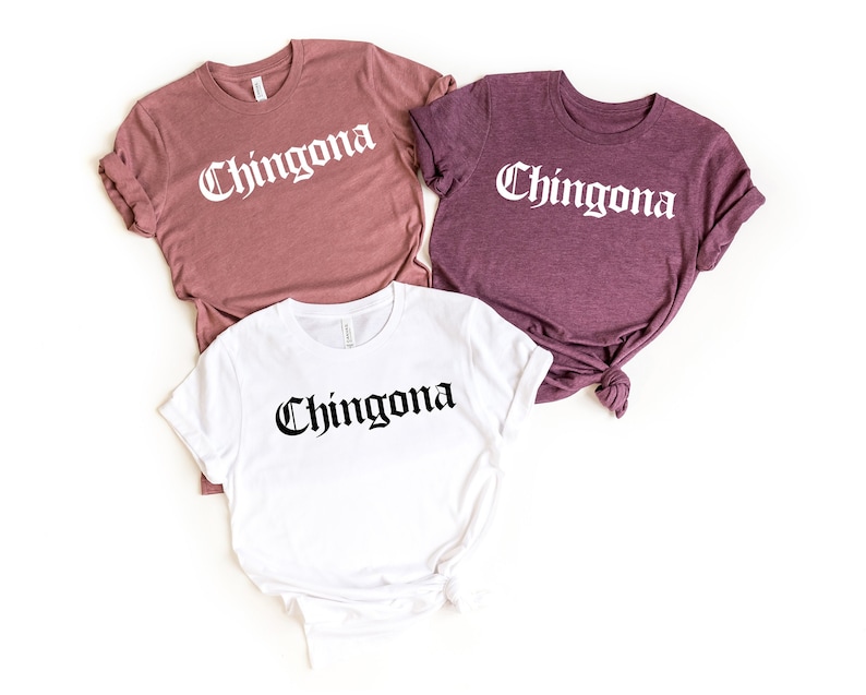 May include: Three t-shirts with the word "Chingona" printed in black gothic font on a white background. The shirts are in different colors: mauve, white, and heather purple.