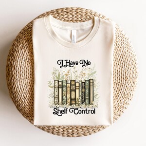 May include: Cream-colored t-shirt with the text "I Have No Shelf Control" above a graphic of vintage books and floral accents. The design features a collection of antique books in various colors, surrounded by delicate botanical illustrations.