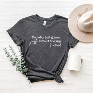 Puede incluir: Camiseta gris oscuro con el texto "Probably just gonna jingle some of the way. I'm tired." Un sombrero beige y una taza blanca est&aacute;n junto a la camiseta. Un manojo de tallos de eucalipto falsos verdes y grises tambi&eacute;n est&aacute; en la imagen.