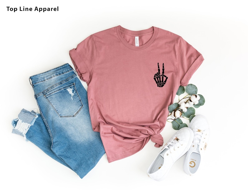 May include: A mauve t-shirt with a black skeleton hand making a peace sign. The t-shirt is paired with blue jeans and white sneakers.