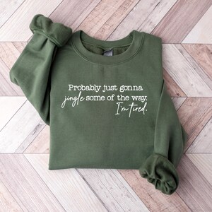 Puede incluir: Una sudadera verde oliva con el texto "Probably just gonna jingle some of the way. I'm tired." en escritura blanca. La sudadera est&aacute; tendida sobre una superficie de madera marr&oacute;n claro y blanca.