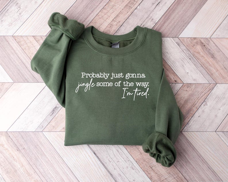 Puede incluir: Una sudadera verde oliva con el texto "Probably just gonna jingle some of the way. I'm tired." en escritura blanca. La sudadera est&aacute; tendida sobre una superficie de madera marr&oacute;n claro y blanca.