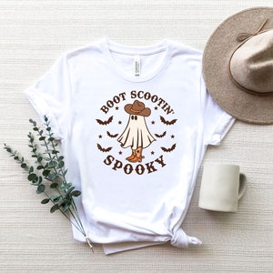 May include: White t-shirt with a ghost wearing cowboy boots and a hat. The text on the shirt reads "Boot Scootin Spooky".