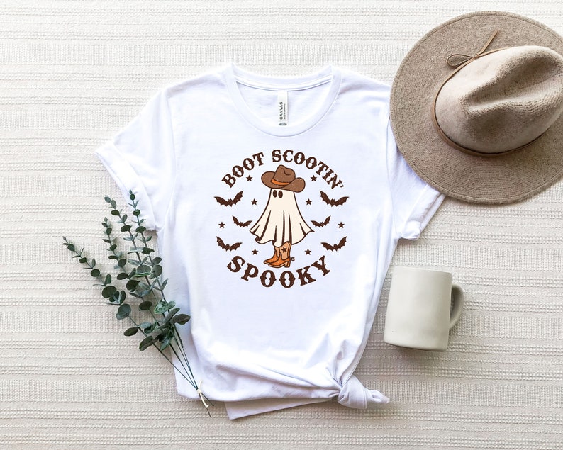 May include: White t-shirt with a ghost wearing cowboy boots and a hat. The text on the shirt reads "Boot Scootin Spooky".