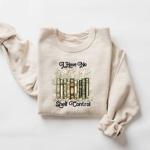 May include: Beige sweatshirt with the text "I Have No Shelf Control" in black script. The graphic features a stack of books with floral accents. The sweatshirt is made of a soft material and has long sleeves.