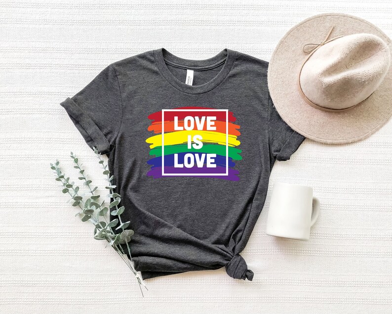 May include: Dark gray t-shirt with the text "LOVE IS LOVE" in a white outlined box, set against a rainbow-colored brushstroke design. A neutral-toned hat and white mug are next to the shirt.