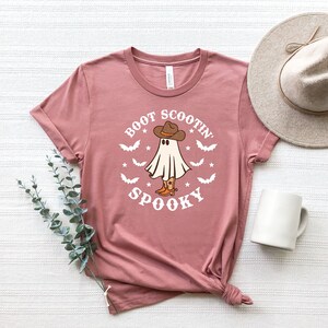May include: A dusty rose t-shirt with a white graphic of a ghost wearing cowboy boots and a cowboy hat. The text "Boot Scootin' Spooky" is printed on the shirt.
