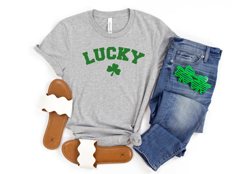 May include: A gray t-shirt with the word "LUCKY" in green letters and a green shamrock underneath. The shirt is paired with blue jeans and white sandals.
