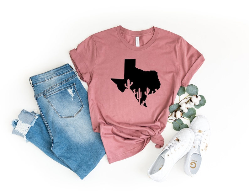 May include: A mauve t-shirt with a black silhouette of the state of Texas with cacti inside. The t-shirt is laying on a white surface with a pair of blue jeans, white sneakers, and a sprig of eucalyptus.