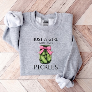 Puede incluir: Sudadera gris con el texto "Just a girl who loves pickles" y un gr&aacute;fico de un frasco de pepinillos con un lazo rosa.