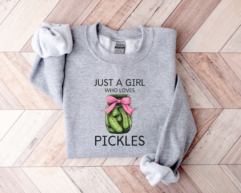 Puede incluir: Sudadera gris con el texto "Just a girl who loves pickles" y un gr&aacute;fico de un frasco de pepinillos con un lazo rosa.